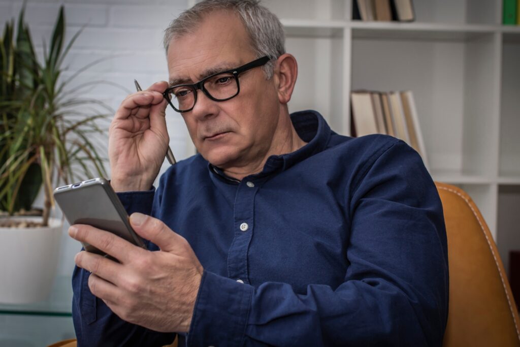 Signore brizzolato seduto su una poltrona mentre guarda il cellulare con gli occhiali da lettura